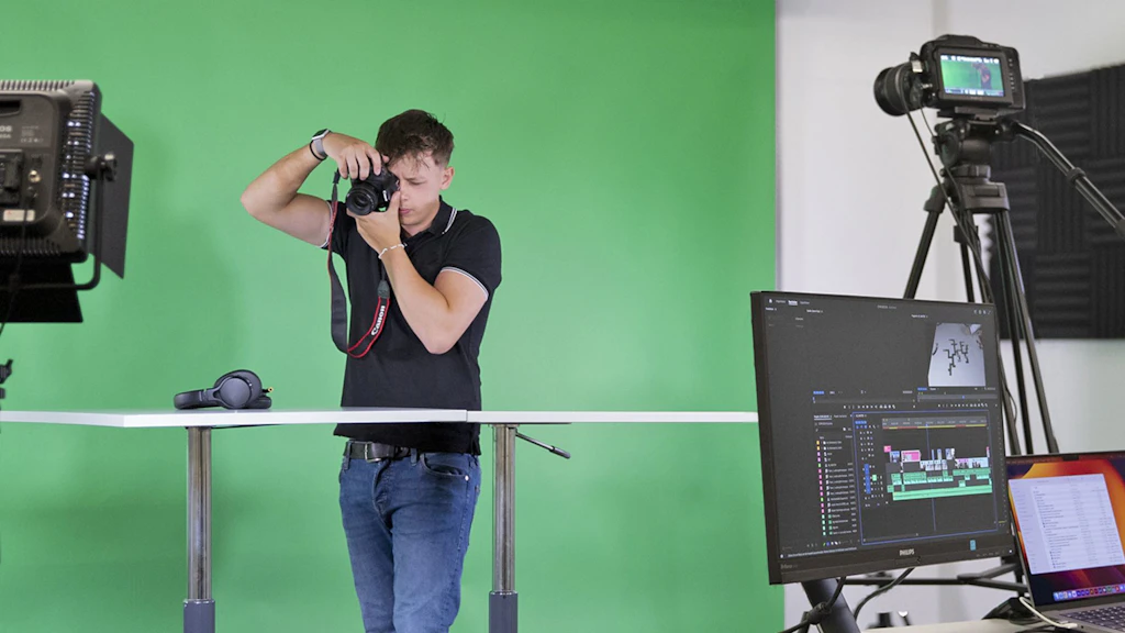 Person mit Kamera vor grünem Hintergrund in einem Fotostudio mit Monitoren und Beleuchtung
