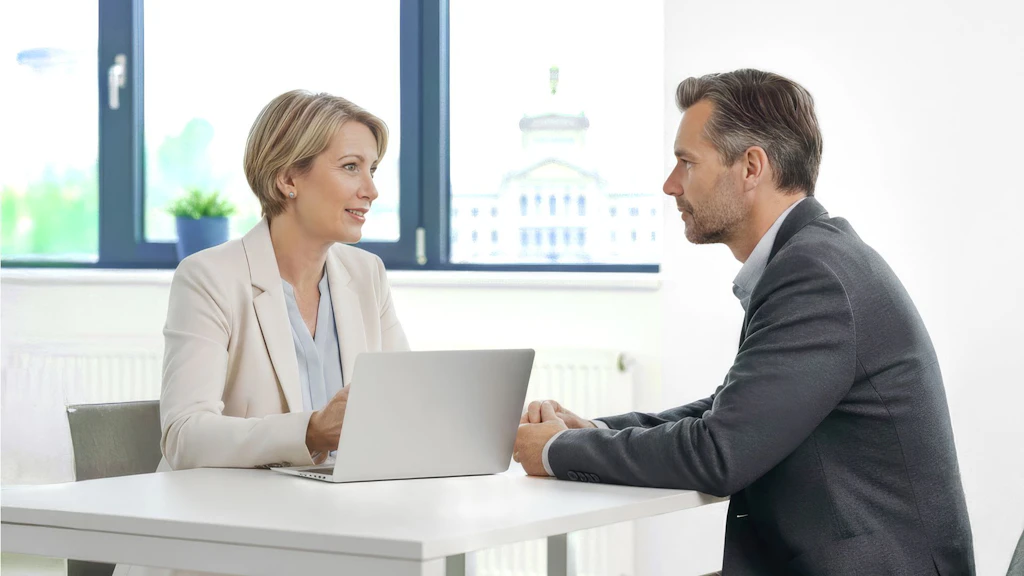Zwei Personen sitzen sich an einem weißen Tisch in einem hellen Büro gegenüber und führen ein Gespräch