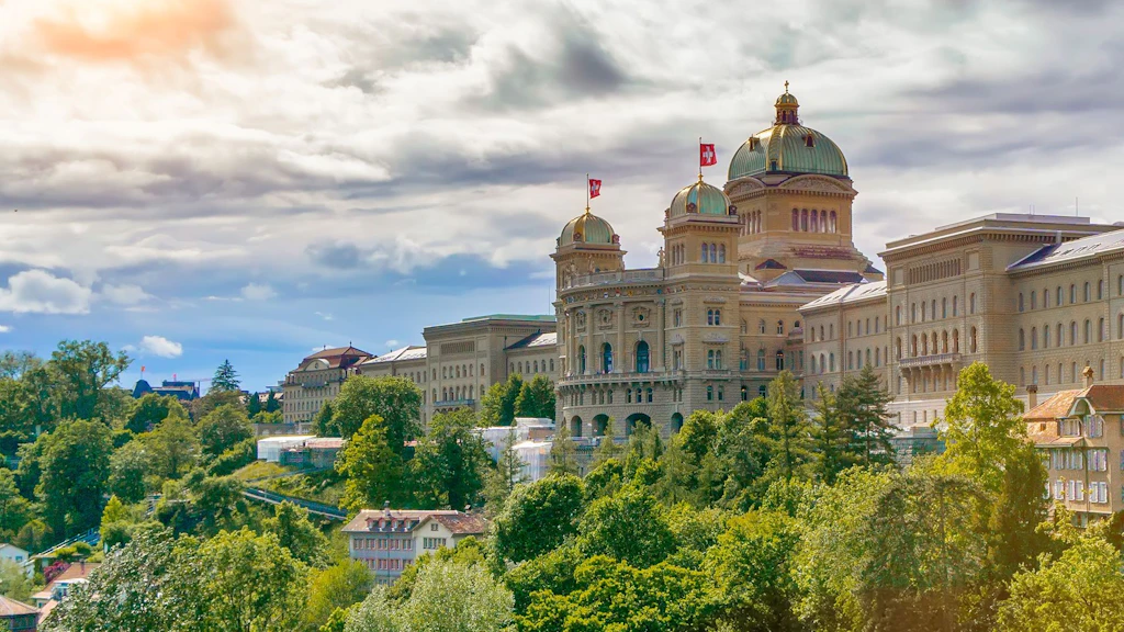 Bundeshaus in Bern mit Kuppeln und Schweizer Fahnen umgeben von Bäumen und Gebäuden unter bewölktem Himmel