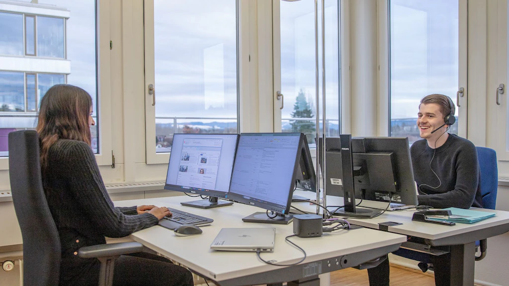 Deux personnes assises à des bureaux opposés avec plusieurs écrans dans un bureau lumineux aux grandes fenêtres