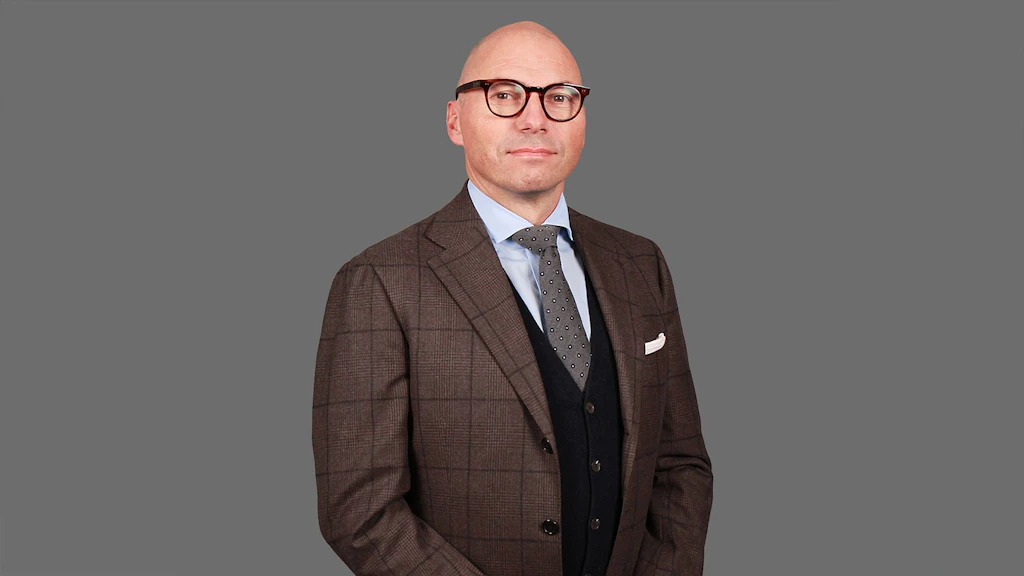 Uomo con giacca marrone a quadri, camicia azzurro chiaro, cravatta a motivi e gilet nero su sfondo grigio